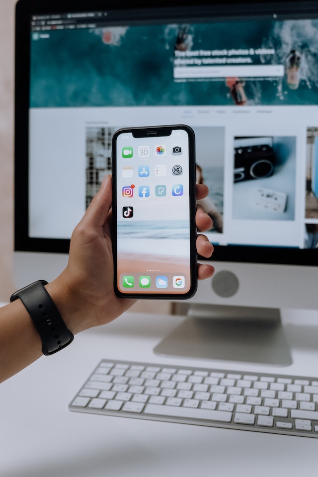 A hand holding a smartphone in front of an Imac.