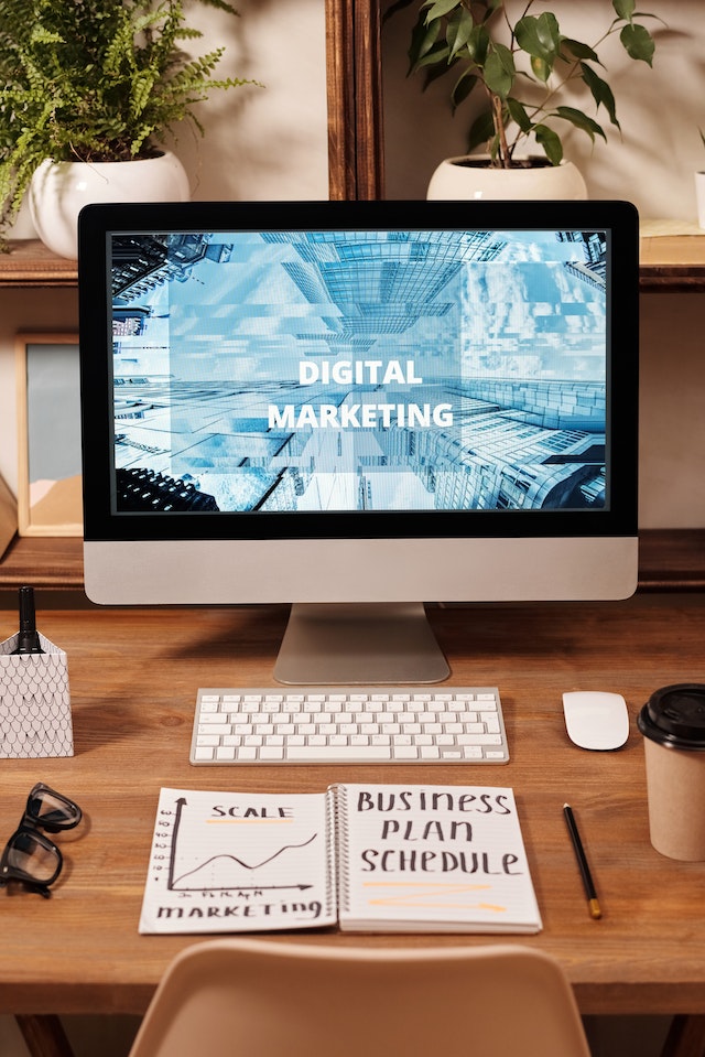 Computer screen displaying a digital marketing graphic in a home office with a notebook showing notes for a business plan.