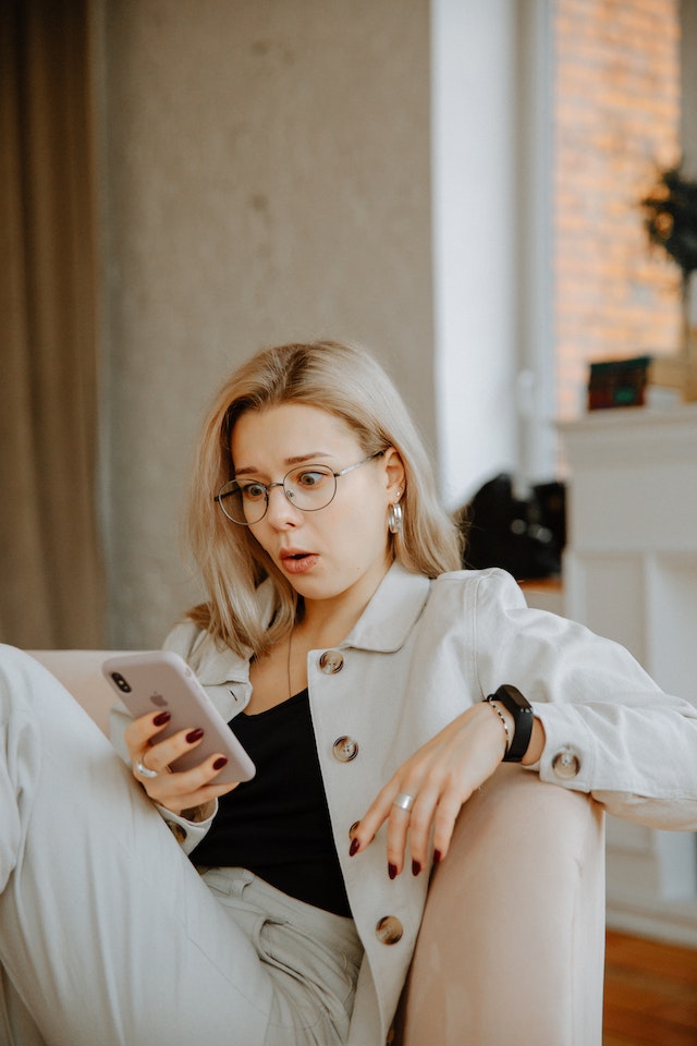 Woman in a beige coat surprised while looking at NSFW content on her smart phone.