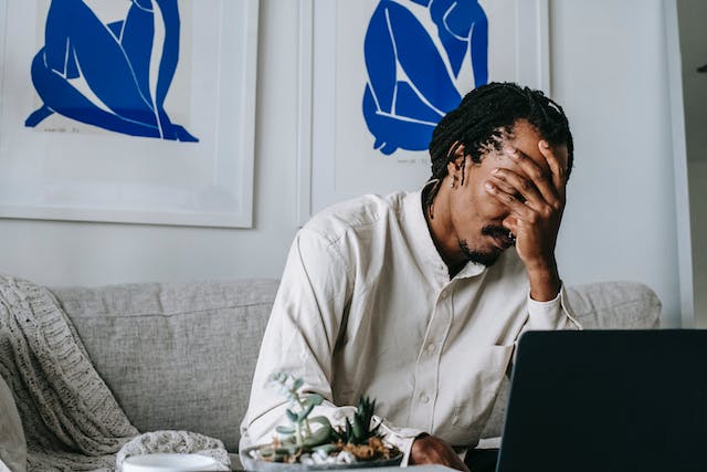 A black man with hands on his face working on a laptop. 