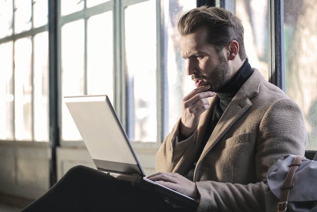 A tense man in a brown jacket is looking at his laptop.