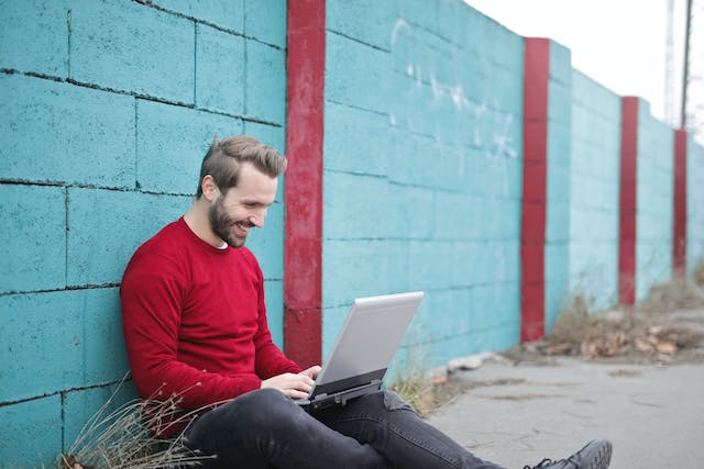 Bărbat în cămașă roșie sprijinit de un perete și folosind un laptop pentru a trimite mesaje pe Instagram.