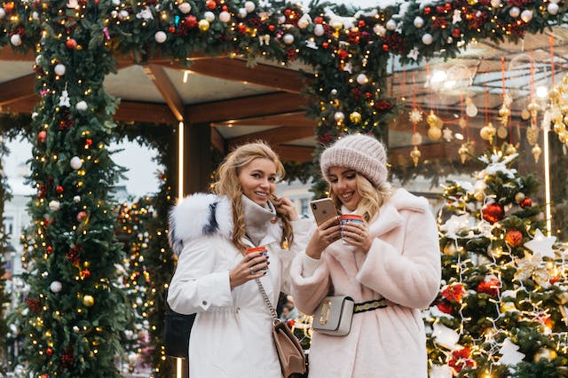 Two female friends in large coats drink coffee together.