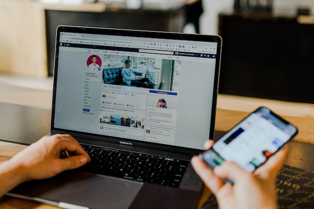 A picture of someone browsing a Facebook profile on a black laptop while holding a smartphone.