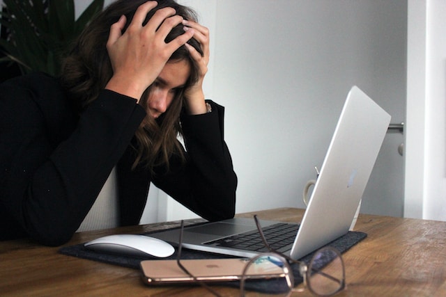 Een gestreste vrouw in een zwarte jurk die haar hoofd vasthoudt met haar laptop open.