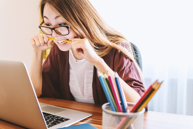 A woman in glasses is biting a pencil and looking at some blocked accounts on her computer. 