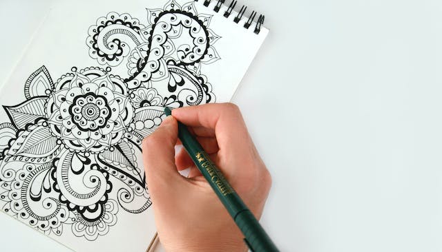 Alguém a desenhar rabiscos complexos de uma flor num caderno.