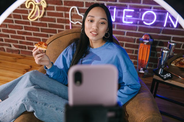 A female influencer eating a slice of pizza while filming a vertical video with her iPhone and tripod.