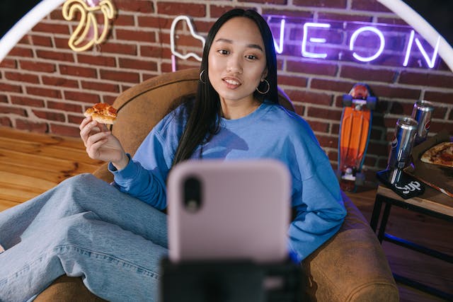 Una influencer che mangia un trancio di pizza mentre gira un video verticale con l'iPhone e il treppiede.