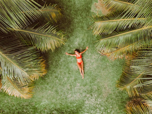 A travel influencer trees in her swimwear relaxing on the grass next to palm trees.