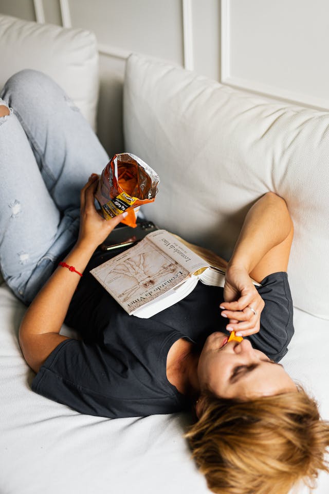 A woman carelessly lying in bed while eating chips.