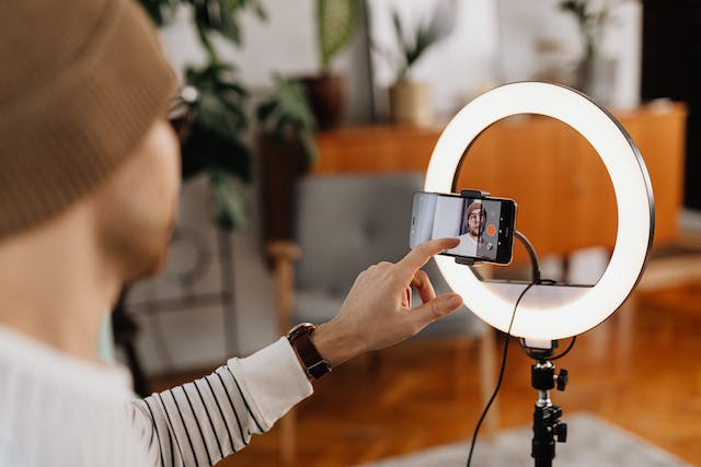 Een mannelijke maker filmt een video van zichzelf met zijn telefooncamera, een statief en een ringlamp.