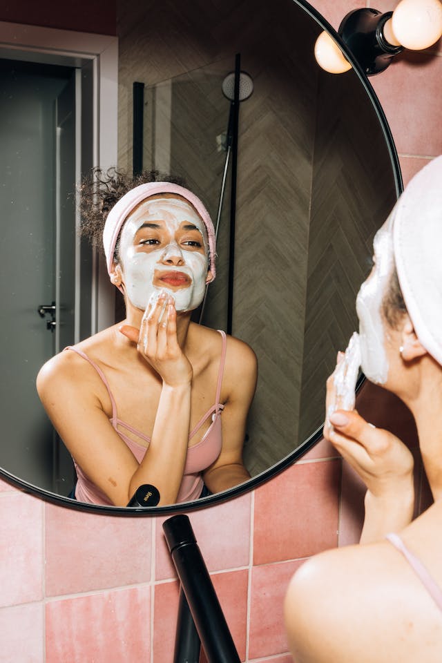 Une femme aux cheveux relevés applique un masque facial devant le miroir de la salle de bains.