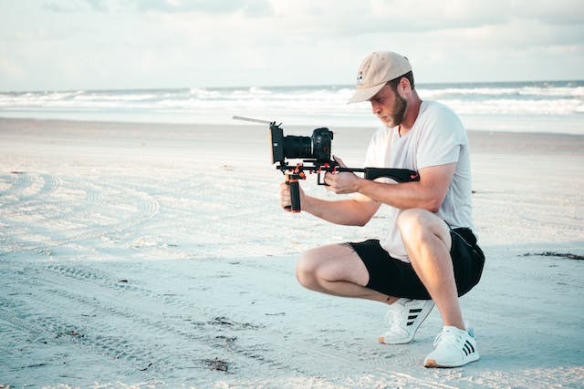 Un influencer care ține în mână și înregistrează cu un aparat foto DSLR la malul mării.