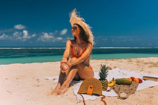 A female influencer sitting on a beach with tropical fruits and a rattan bag on her mat.