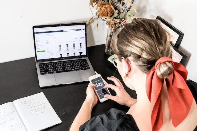 Una mujer editando su historia Instagram en su iPhone con su portátil abierto delante de ella.