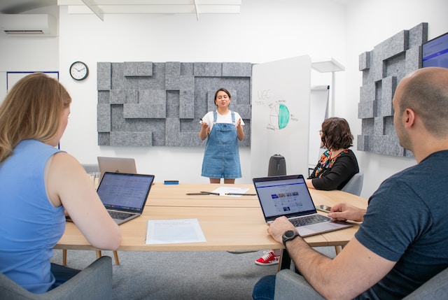 A group sits around a table with laptops, listening to a woman wearing a blue Dungaree.