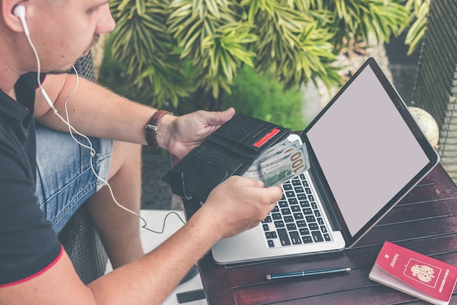 Een persoon die zijn portemonnee met biljetten van 100 dollar voor zijn laptop en paspoort houdt.