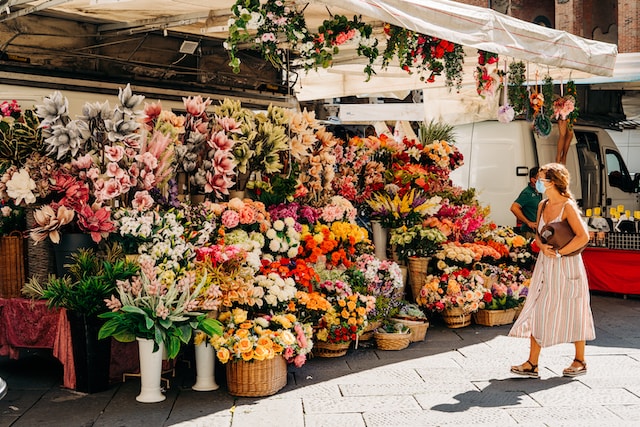 امرأة ترتدي قناعا للوجه تنظر إلى الزهور في كشك سوق محلي وهي تمشي بجانبها.