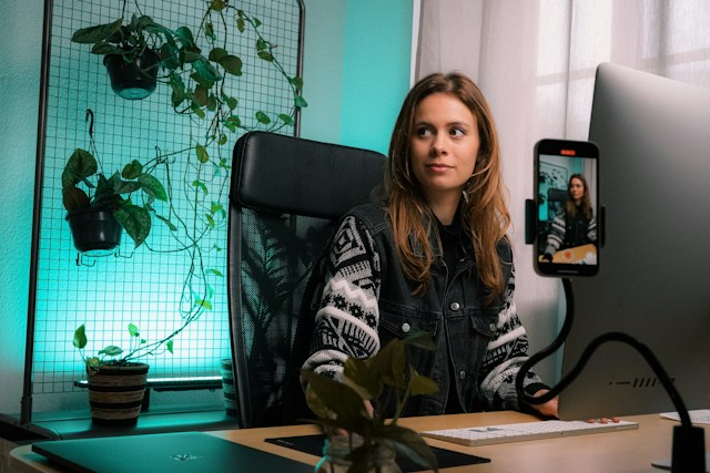 Uma mulher está sentada em frente ao seu computador, a gravar um Reel no seu telemóvel.