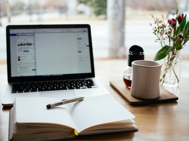 Una instantánea de un cuaderno abierto con un bolígrafo y un MacBook Pro de fondo. 