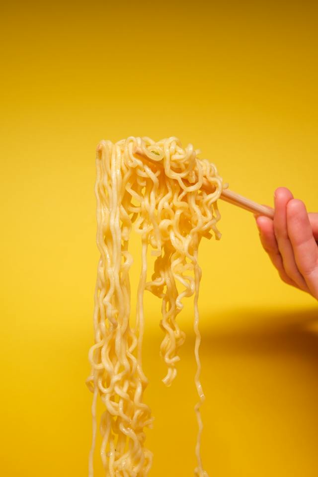 Uma mão a segurar uns noodles instantâneos cozinhados com um par de pauzinhos.