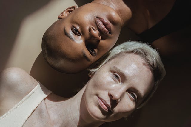 A light-skinned woman and a dark-skinned woman lying on the floor with their heads side-by-side.