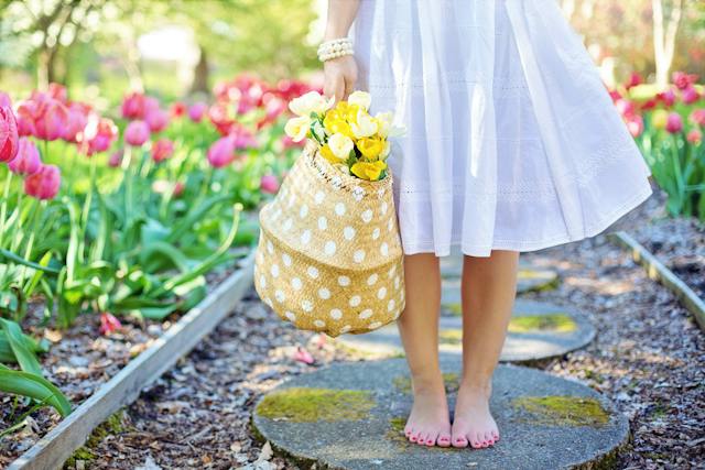 Una donna in abito bianco con in mano un cesto di fiori gialli in un giardino di tulipani.