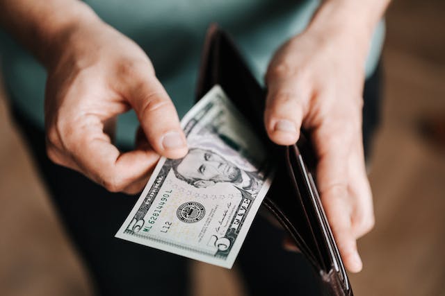 Un bărbat își întinde portofelul din piele maro și scoate o bancnotă de cinci dolari.