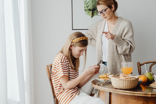 A stressed-looking mother watching her young daughter scroll through social media on a cellphone.