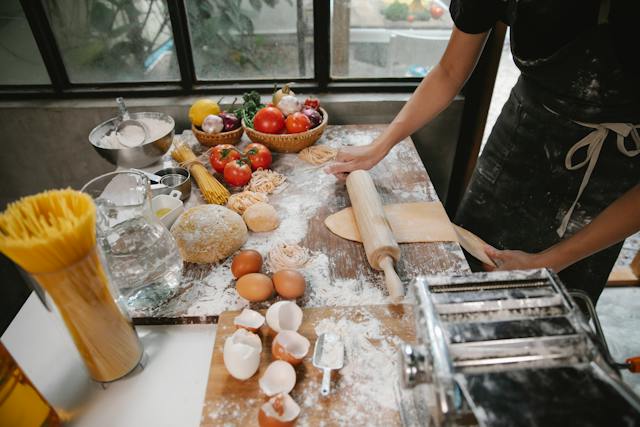 A messy, crowded kitchen counter with many pasta ingredients and kitchen tools.
