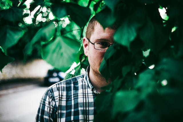 An adult man in a plaid shirt hiding behind the leaves of a tree.