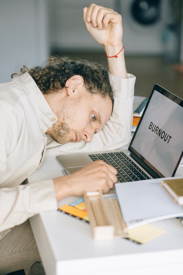 Um empregado cansado descansa a cabeça numa secretária em frente ao seu computador portátil, que apresenta a palavra "Burnout".