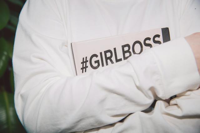 A woman holding a notebook with the hashtag #GIRLBOSS close to her chest.