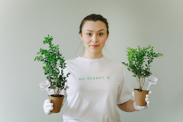 O femeie care ține în mână niște plante proaspăt ghivecate, purtând un tricou pe care scrie "No Planet B."