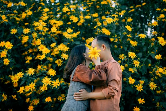 Un hombre y una mujer en un jardín de flores utilizando una flor amarilla para ocultar su beso.