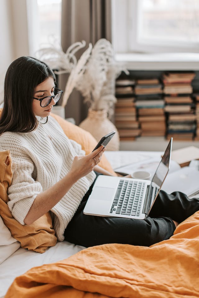 O fată care poartă ochelari caută utilizatorii Instagram pe telefonul și laptopul ei. 