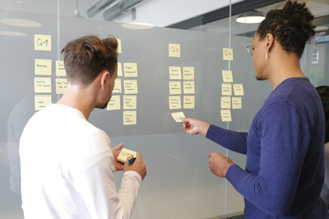 Two men in blue and white shirts are writing on sticky notes and pasting them on a board. 