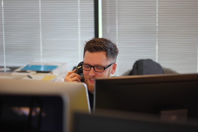 A man in glasses answering a phone call in front of computers.
