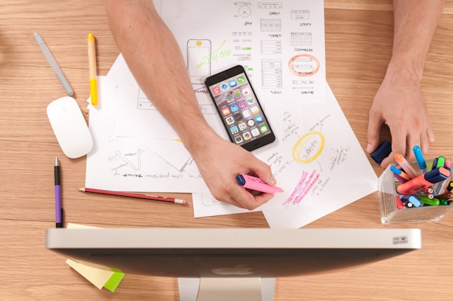 Une personne surligne quelque chose sur une feuille de papier, avec des stylos éparpillés et un téléphone à proximité.