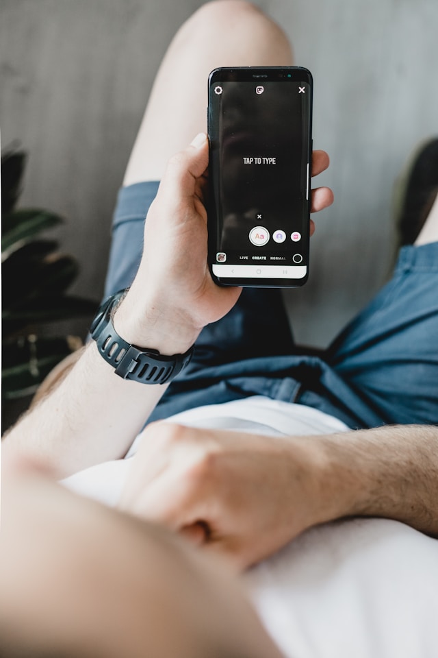 Eine Person in einem weißen Hemd und blauen Shorts hält ein schwarzes Telefon in der Hand und erstellt eine Instagram Story.