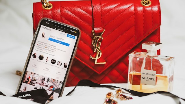 Un telefono che mostra un profilo Instagram accanto a una borsa rossa e a una boccetta di profumo. 