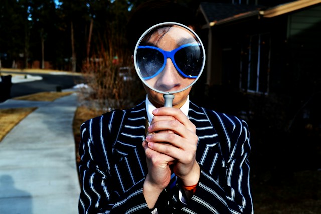 A man holding a magnifying glass in front of his face, enlarging his sunglasses and nose in the process.