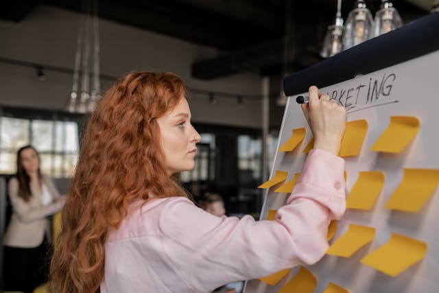 Une femme rédigeant des plans de marketing sur un tableau blanc rempli de notes autocollantes.