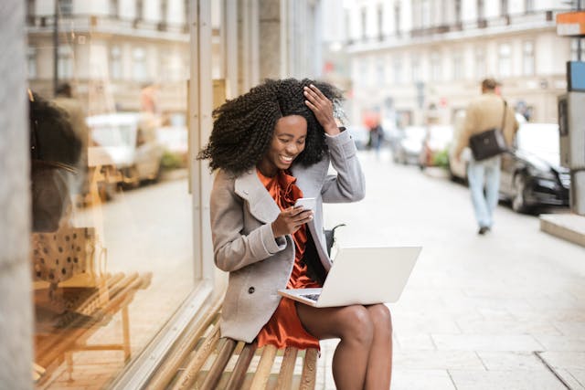 Een vrouw glimlacht terwijl ze haar telefoon doorneemt voor een glazen raam.