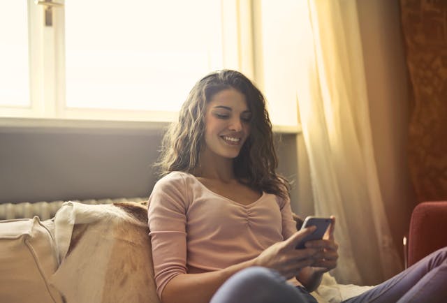 O femeie fericită stând pe pat în timp ce își folosește telefonul.