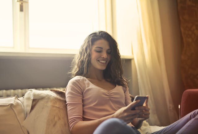 Een gelukkige vrouw zit op bed terwijl ze haar telefoon gebruikt.