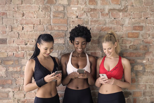 Drie fitnessminnende vrouwen glimlachen terwijl ze naar hun telefoon kijken.