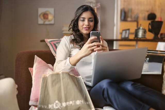 Une femme navigue sur son téléphone avec un ordinateur portable sur les genoux.