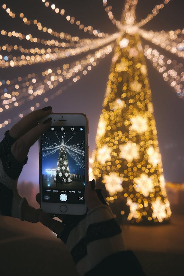 스마트폰으로 불이 켜진 크리스마스 트리 사진을 찍는 사람.